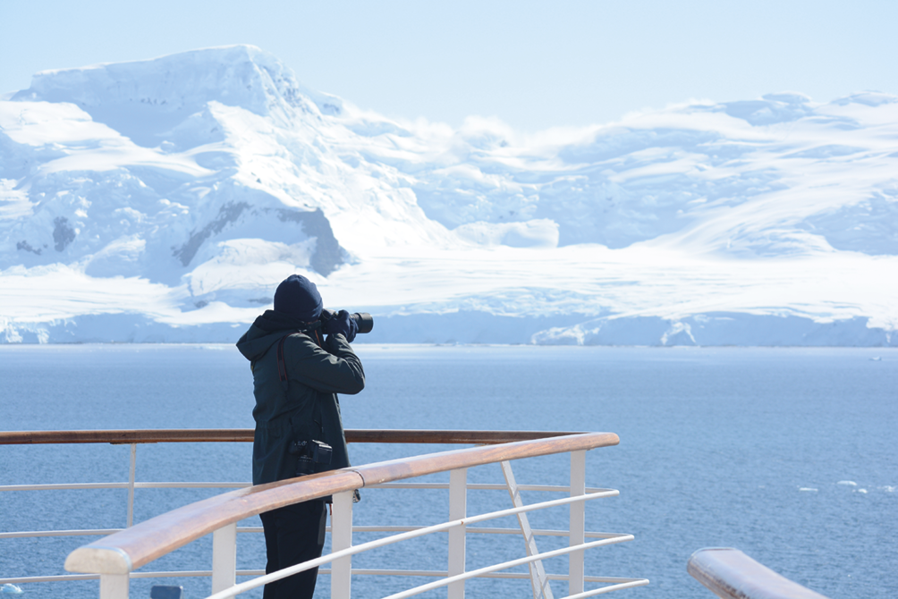 Fotografia. Destaque para um homem em pé em frente ao mar e geleiras. Ele aponta uma máquina fotográfica para o horizonte. Há um parapeito gradeado em frente a ele.
