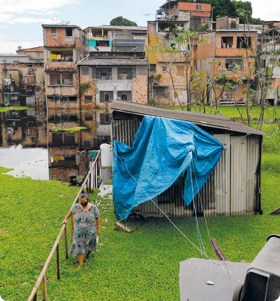 Fotografia. No primeiro plano, uma pequena casa feita com telhas, coberta por uma lona azul em uma área gramada. Há uma mulher caminhando ao lado dela. Atrás da casa há um córrego com casas amontoadas com tijolos aparentes.