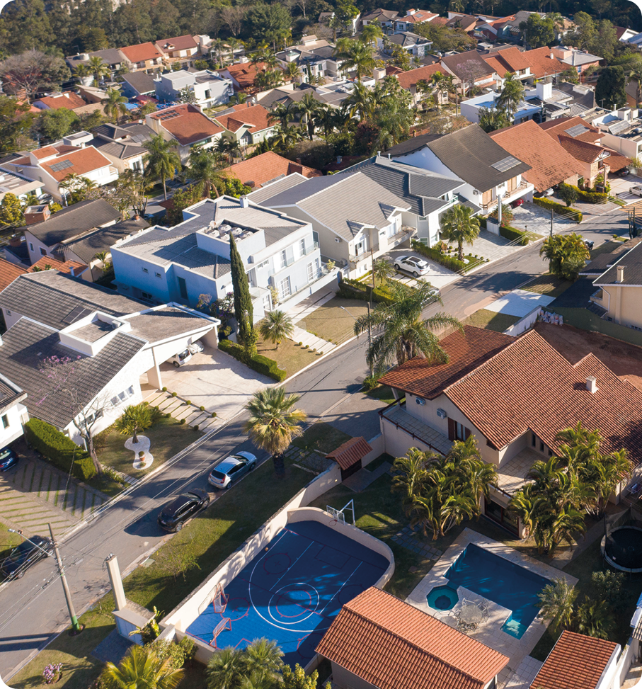 Fotografia. Vista aérea. Bairro com casas grandes. Há uma piscina e uma quadra em uma delas e árvores nas calçadas.