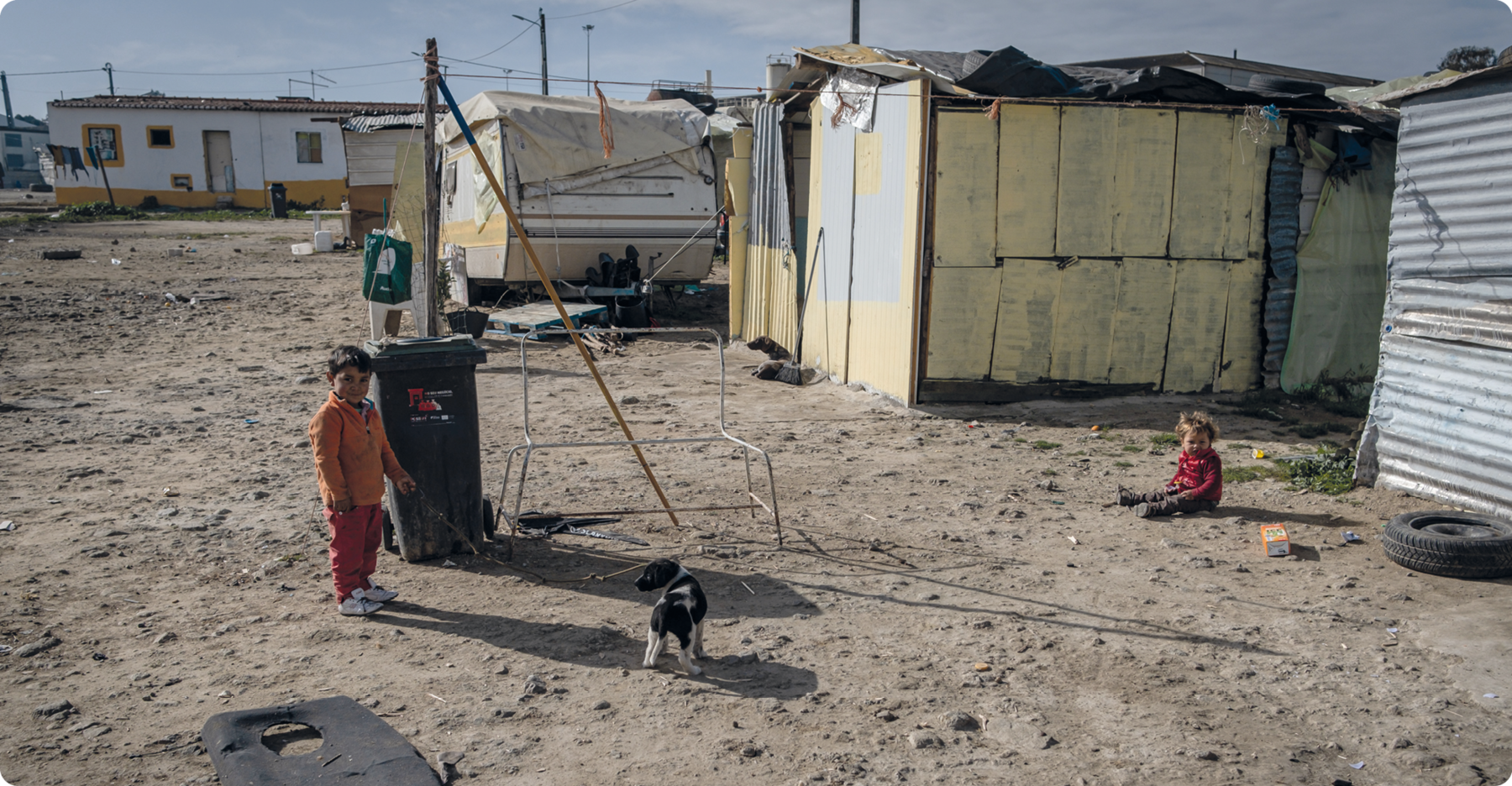 Fotografia. Barracos de tábuas em uma área de terra, ao fundo há dois trailers home. Em frente, uma criança em pé ao lado de um cachorro e outra criança sentada no chão.