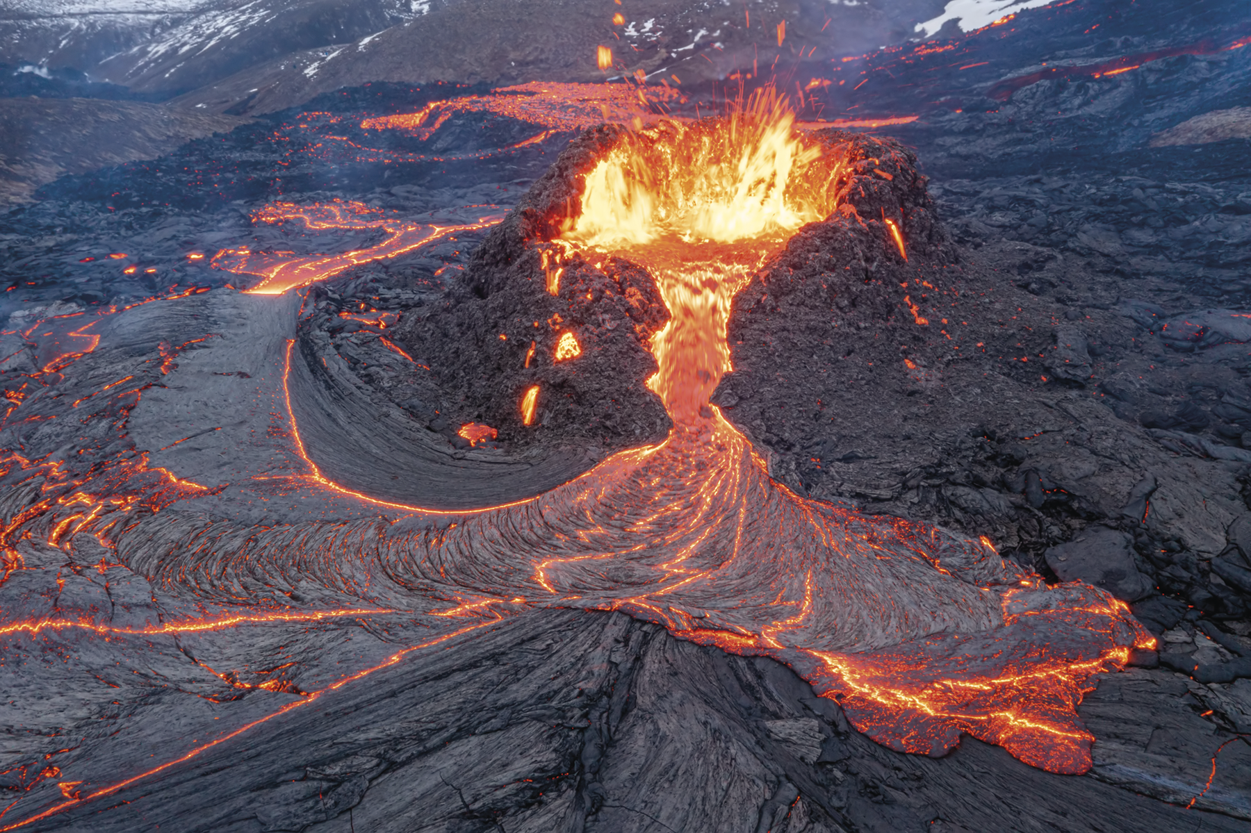 Fotografia. Um vulcão em erupção. Há lava escorrendo e espirrando de sua borda e espalhadas ao redor.