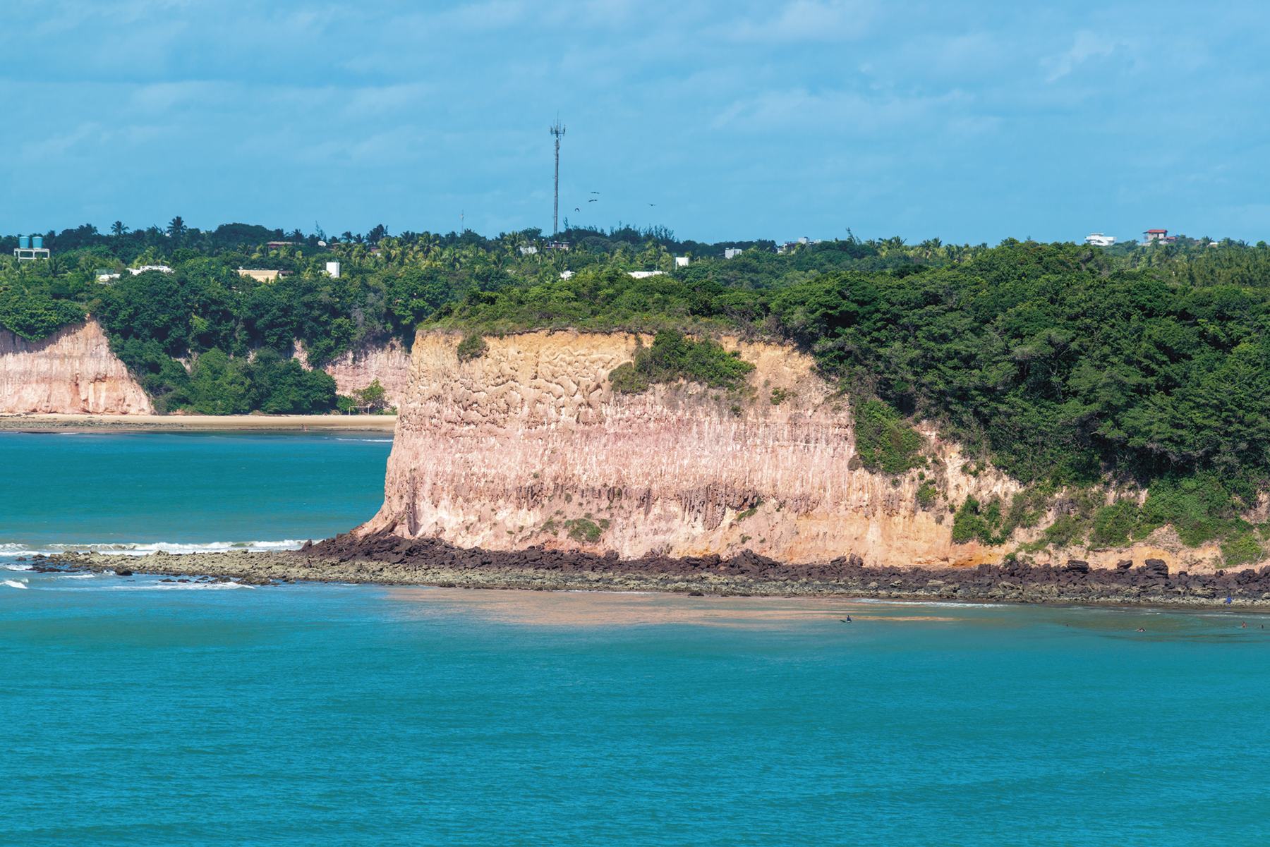 Fotografia. Falésia, formação rochosa de base reta e topo plano, coberta por vegetação, em frente ao mar.