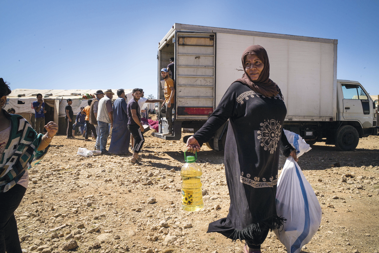 Fotografia. Uma mulher caminhando por uma área de terra. Ela carrega uma garrafa de óleo e um saco. Atrás dela, pessoas enfileiradas em frente a um caminhão.