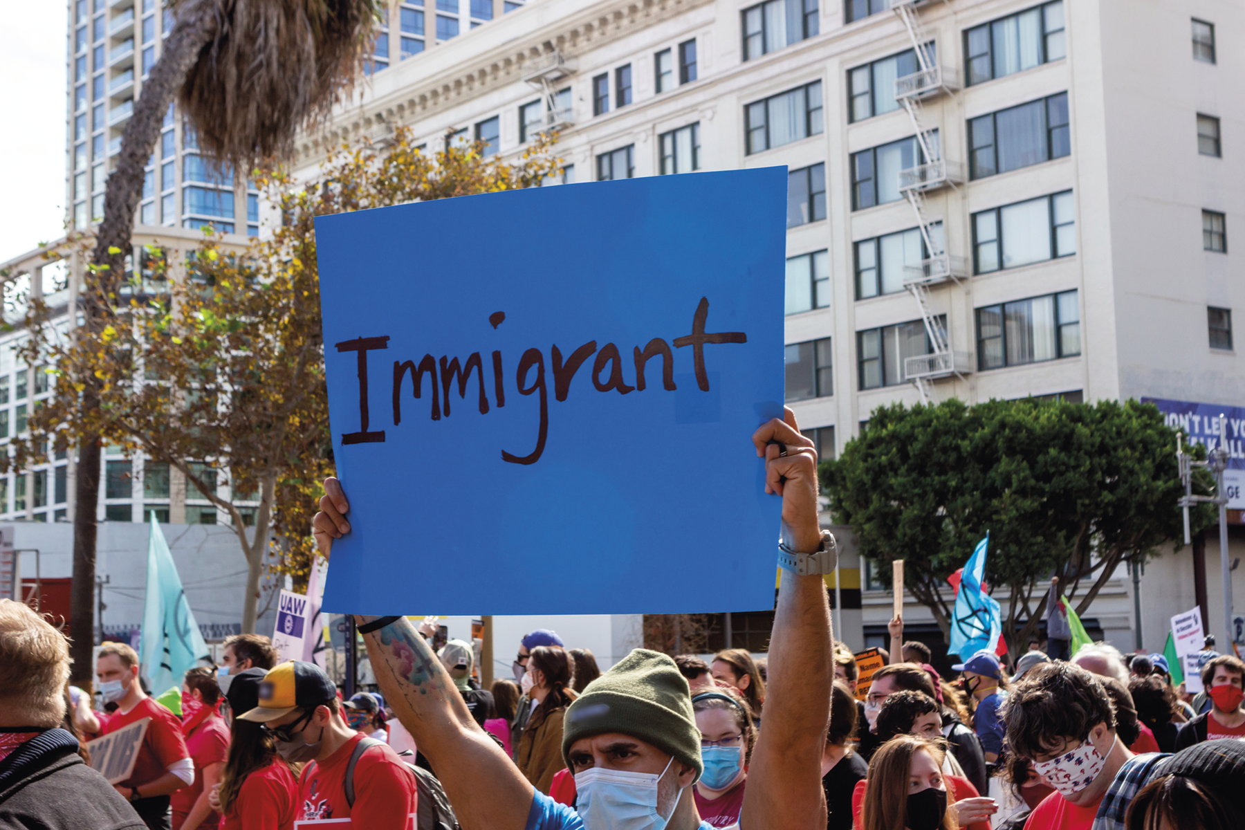 Fotografia. Destaque para uma manifestação repleta de pessoas. Em destaque, um cartaz com a inscrição: Immigrant. Ao fundo, edifícios e algumas árvores.