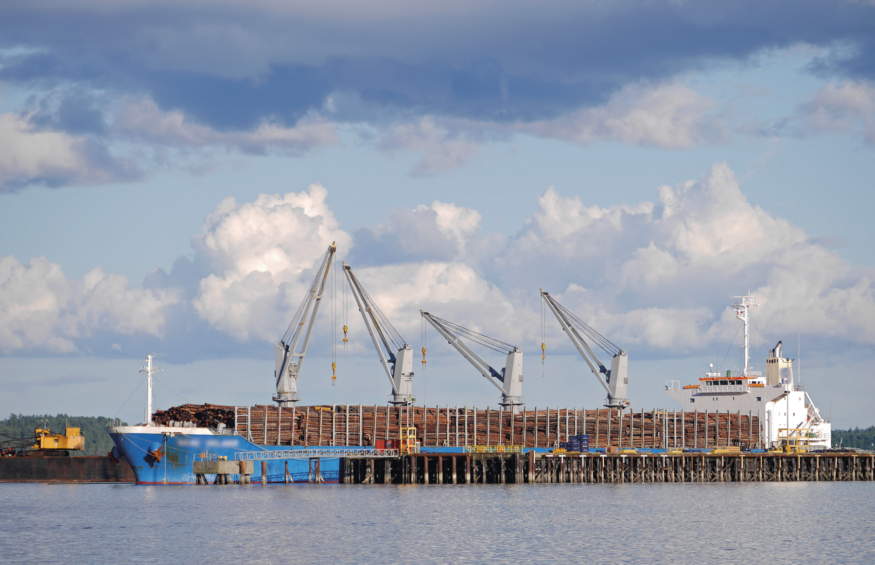 Fotografia. Um navio carregado de troncos de madeira ancorado em um porto. Há gruas manipulando as cargas.