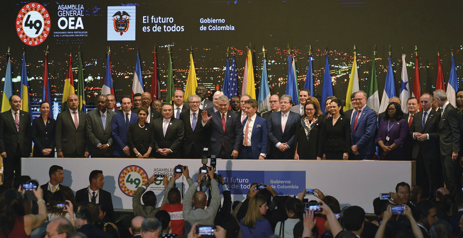 Fotografia. Diversas pessoas reunidas atrás de uma bancada. Todas usam terno e gravatas ou vestidos escuros. Atrás delas há diversas bandeiras hasteadas e um painel com logotipos e foto de uma cidade durante a noite. Em frente a elas há pessoas fotografando.