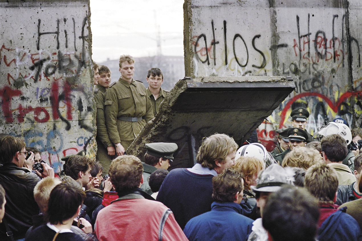 Fotografia. Uma multidão de pessoas reunidas em frente há um muro com pichação. Há uma parte dele sendo retirada, e atrás dela, soldados fardados.