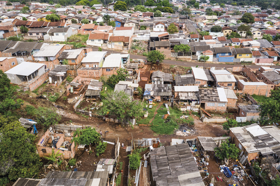Fotografia. Vista aérea. Casas amontoadas em um terreno com um desnível e ruas sem pavimentação. Há árvores entre elas e um terreno com lixo.