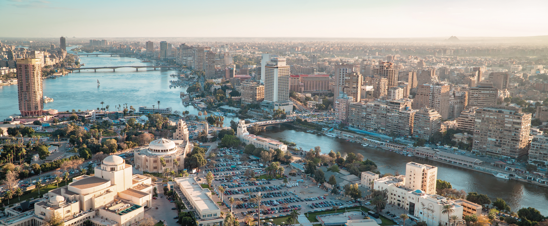 Fotografia. O rio Nilo cortando uma cidade com muitos edifícios. Há diversas pontes sobre ele e na margem esquerda, construções baixas com cúpulas e um grande estacionamento.