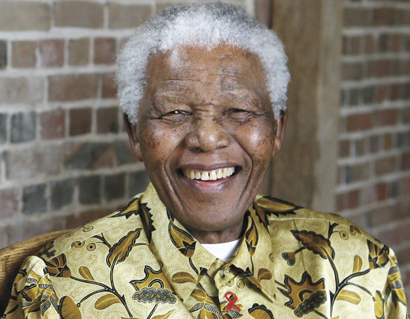 Fotografia. Mandela, homem negro de cabelos curtos, brancos, sorrindo. Ele usa uma camisa estampada.