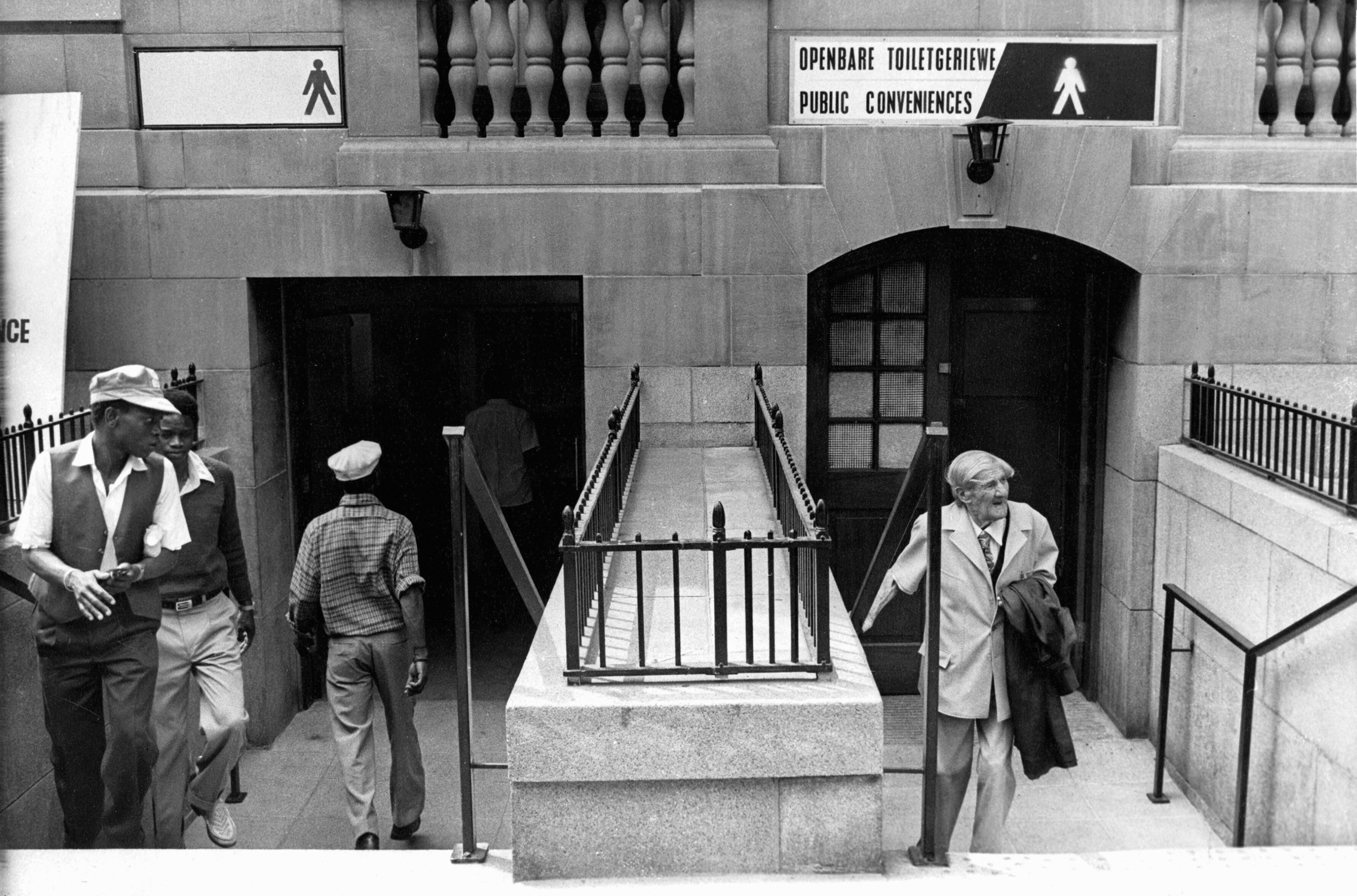 Fotografia em preto e branco. Duas escadas paralelas. Há homens negros passando por uma à esquerda e um senhor branco na escada à direita.