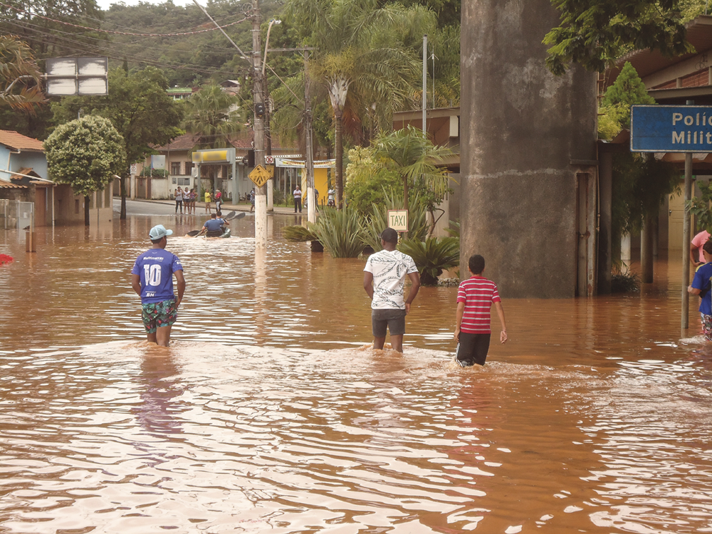 Fotografia. Três pessoas caminhando em uma rua alagada, com a água na altura de seus joelhos. Ao fundo, calçadas com árvores e casas parcialmente submersas.