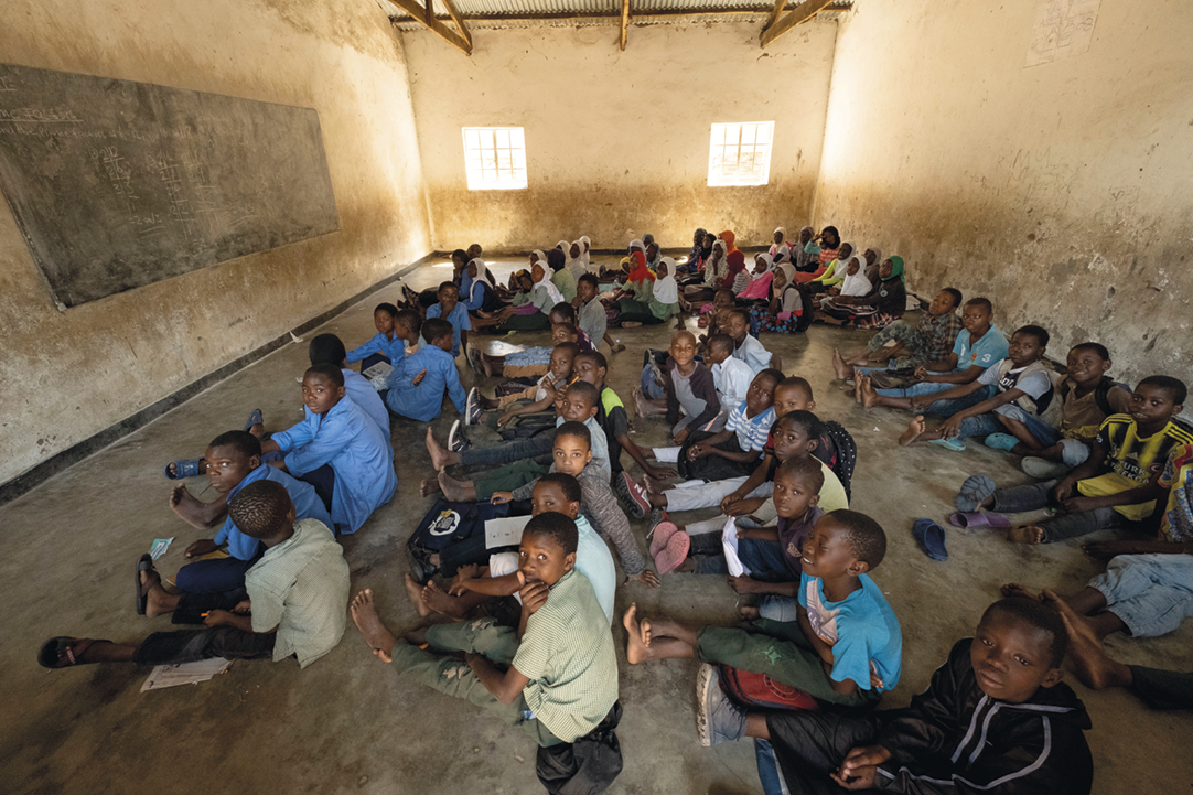 Fotografia. Sala de aula com muitas crianças sentadas no chão, olhando uma lousa que está na parede à esquerda.