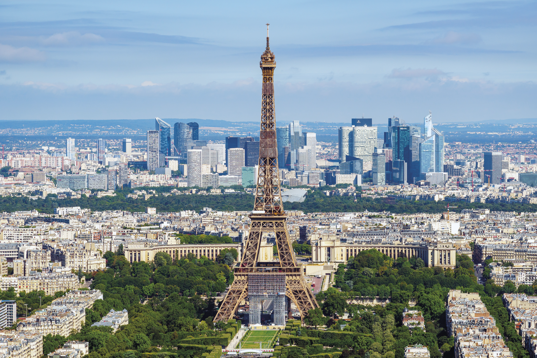 Fotografia. A torre Eiffel com um parque gramado e cercado por árvores. Ao fundo, casas e edifícios.