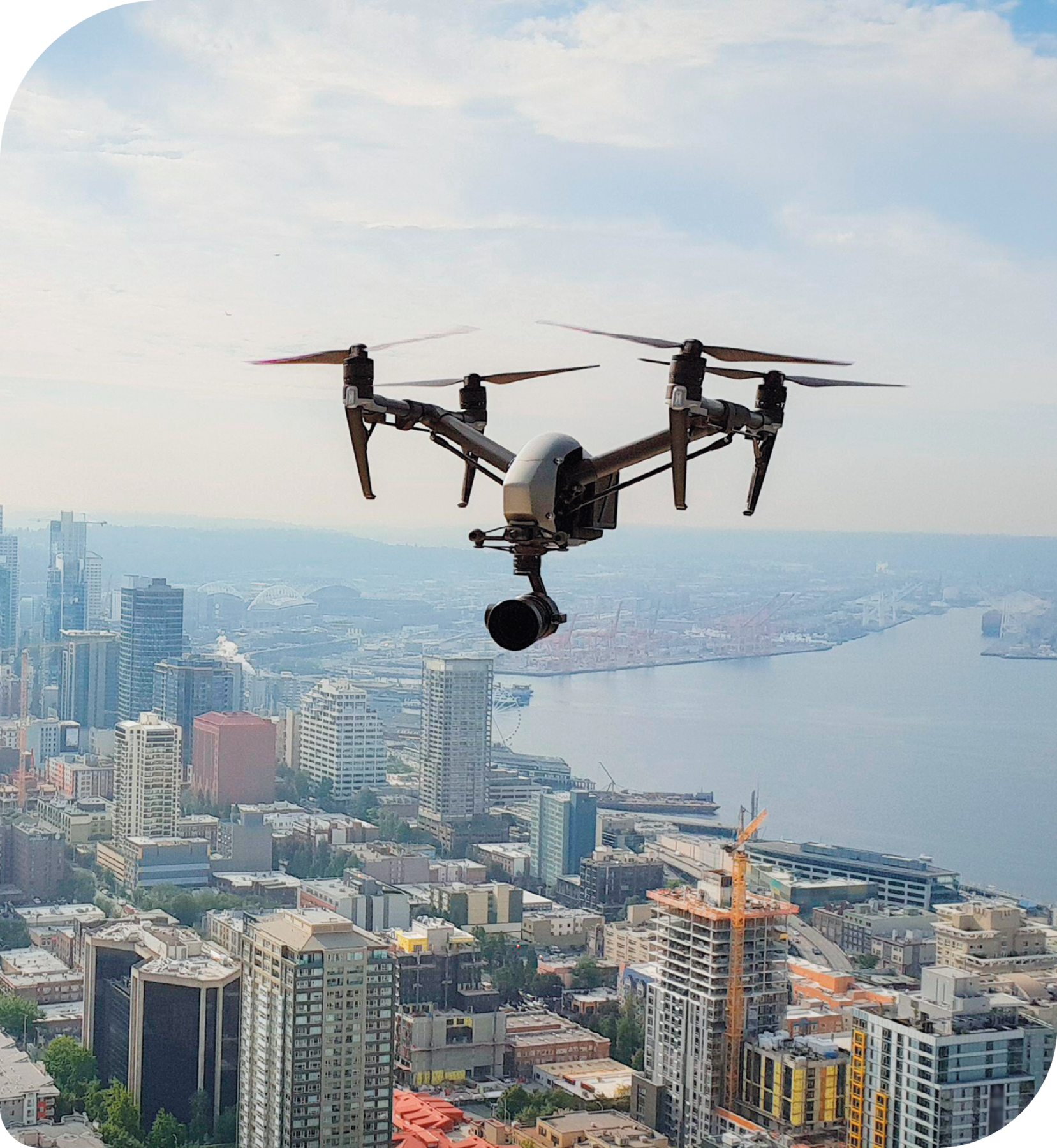 Fotografia. Um drone, equipamento com quatro hélices distintas e uma câmera acoplada na parte inferior. Abaixo dele, uma cidade com diversos edifícios e o mar.