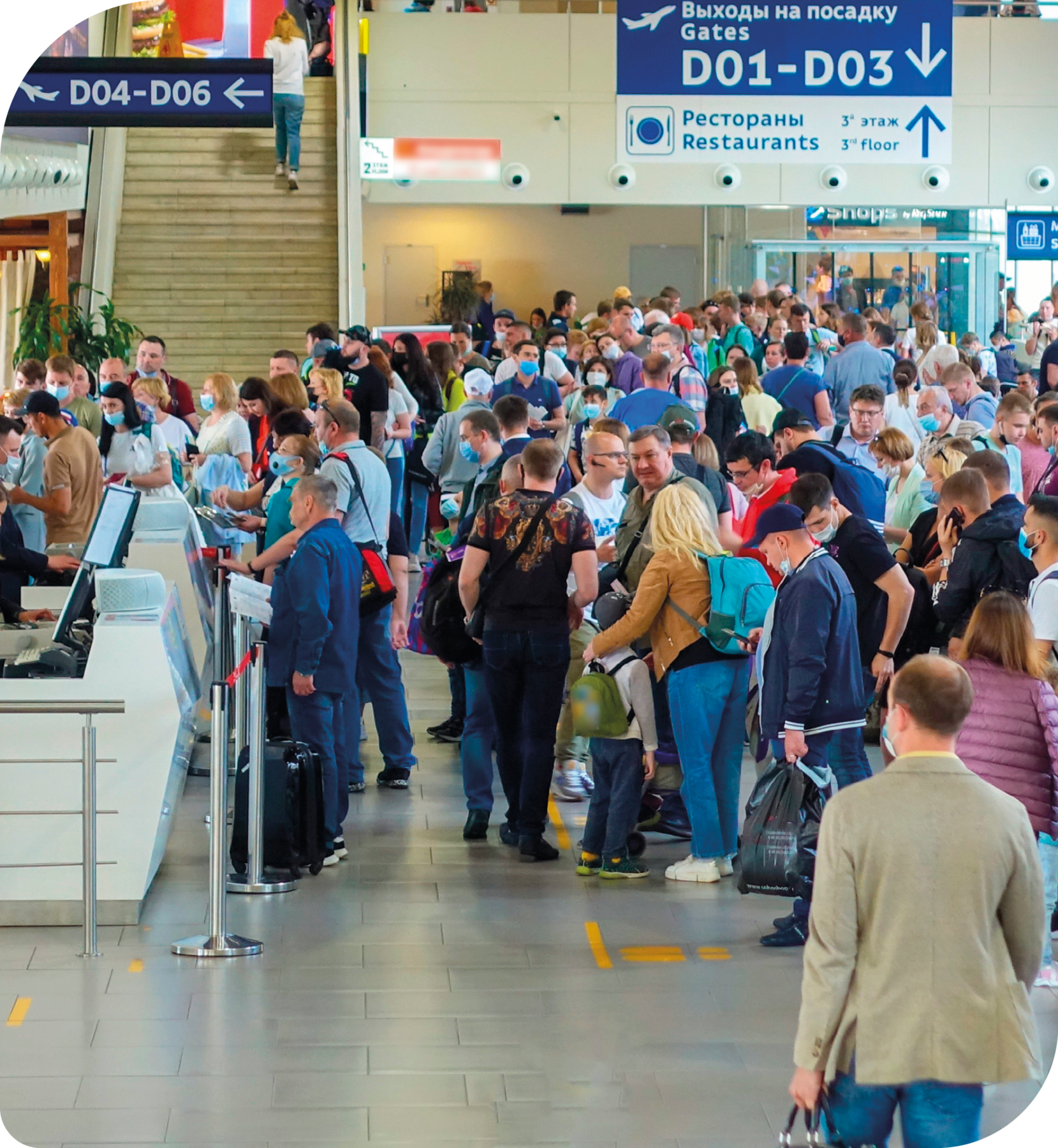Fotografia. Muitas pessoas reunidas em um aeroporto. Eles estão em uma fila em frente a guichês. Ao fundo há placas.