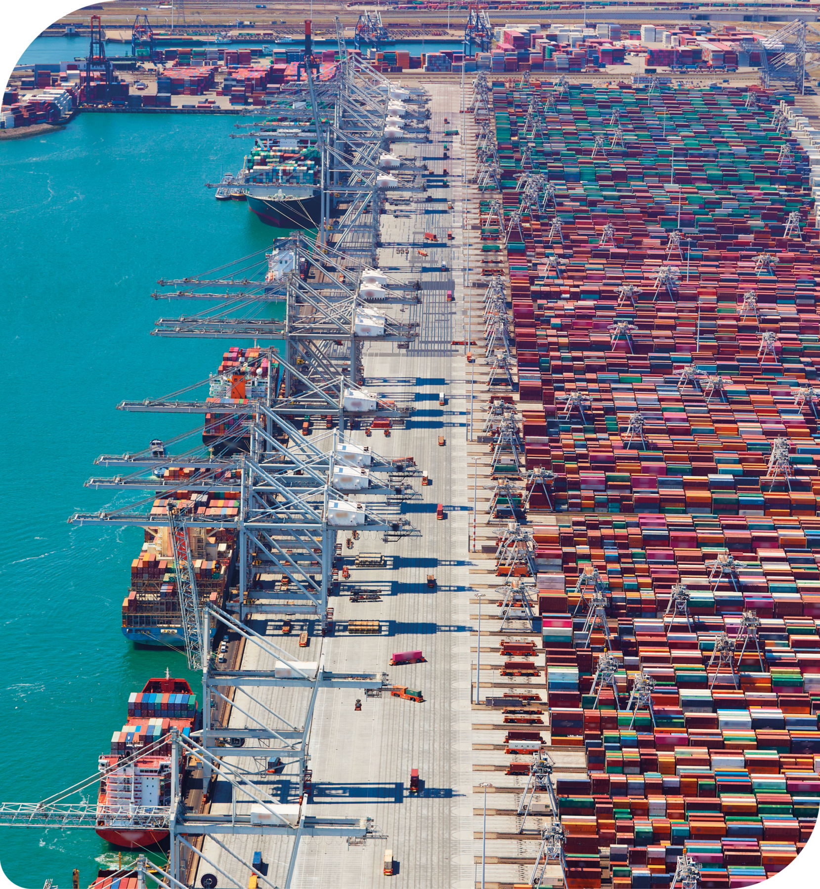 Fotografia. À esquerda, porto com navios ancorados e gruas sobre eles. Ao lado, no porto, pátio com caminhões e à direita, muitos contêineres agrupados.