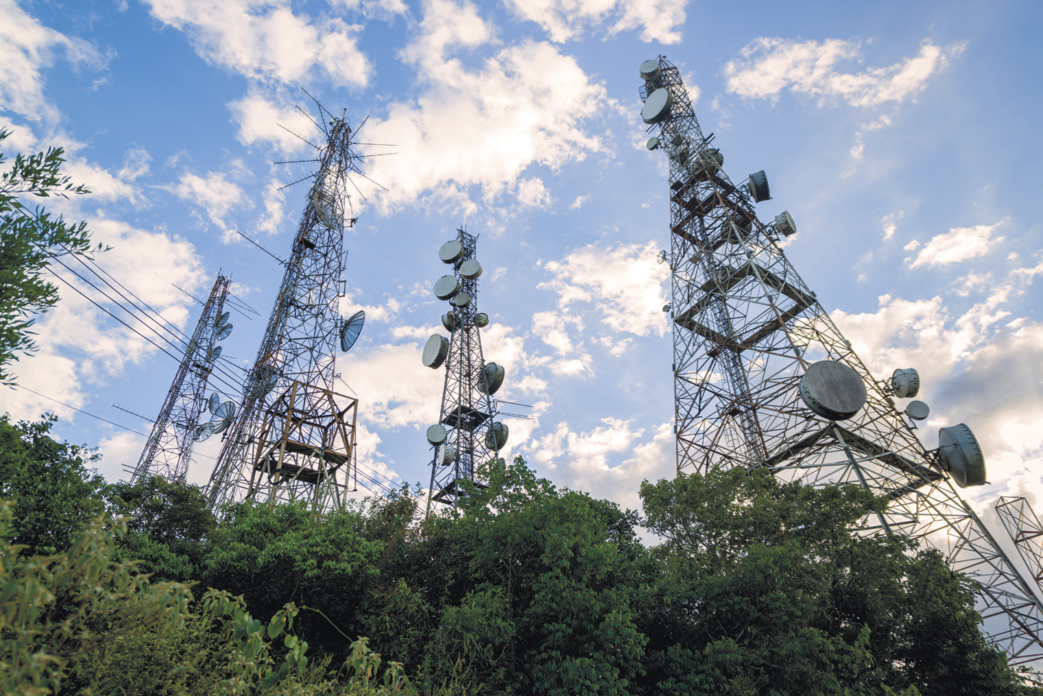 Fotografia. Vista de baixo para cima. Antenas de telecomunicações em meio a árvores. No céu há nuvens.