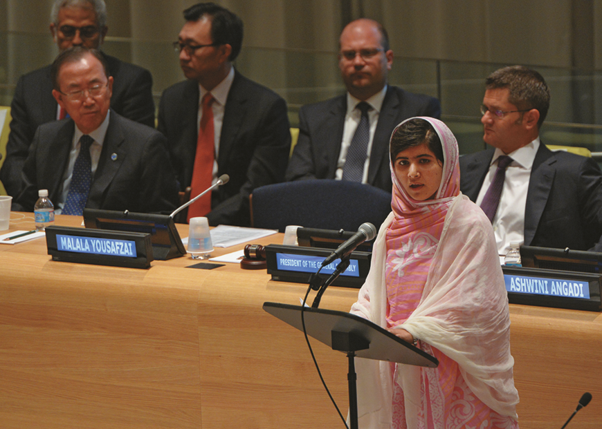 Fotografia. Malala, jovem de roupas claras com um lenço branco cobrindo a cabeça, discursando em frente a um microfone. Atrás dela há homens sentados atrás de uma bancada.