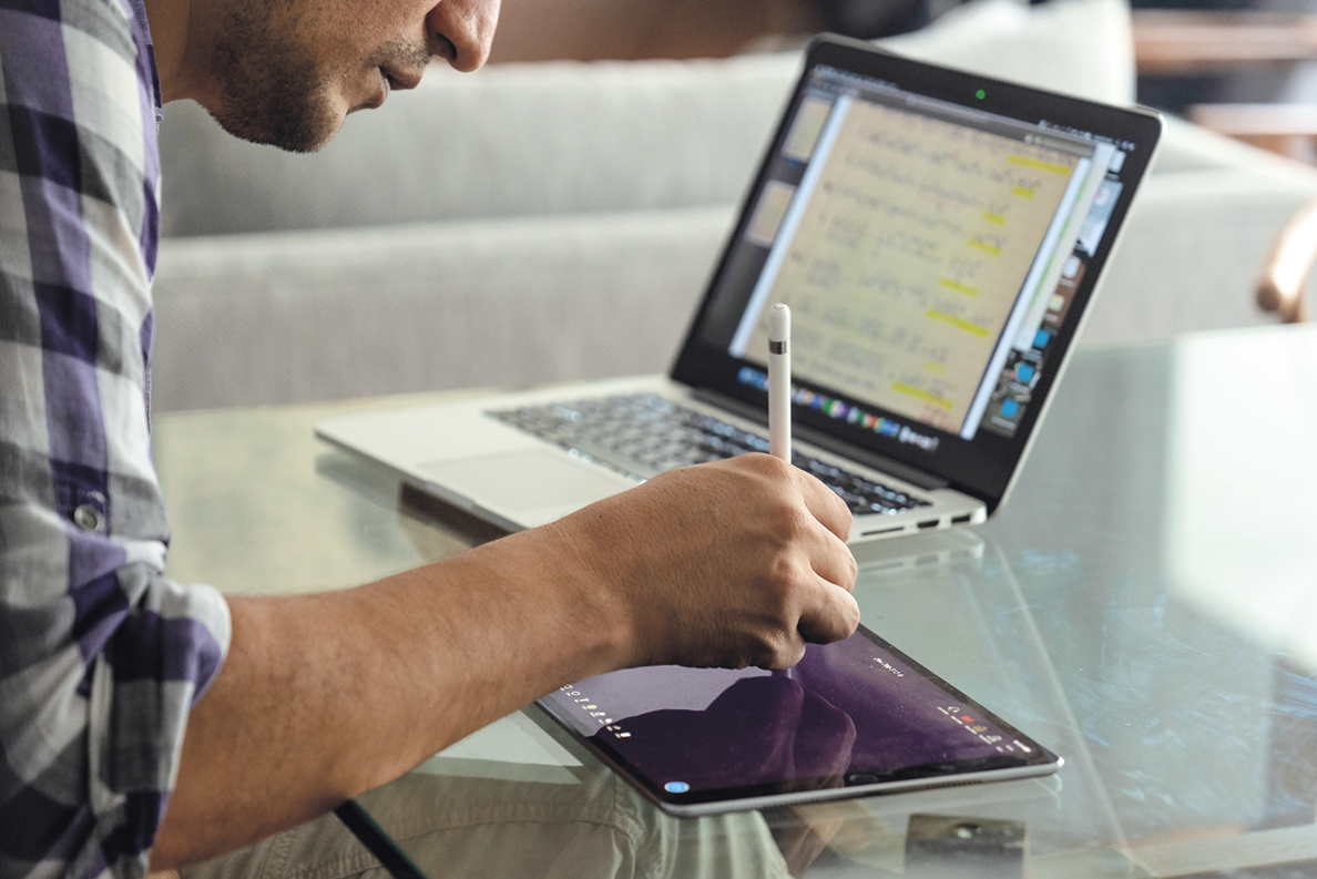 Fotografia. Destaque para um homem utilizando uma espécie de caneta em um tablet. Ao lado dele há um notebook aberto.