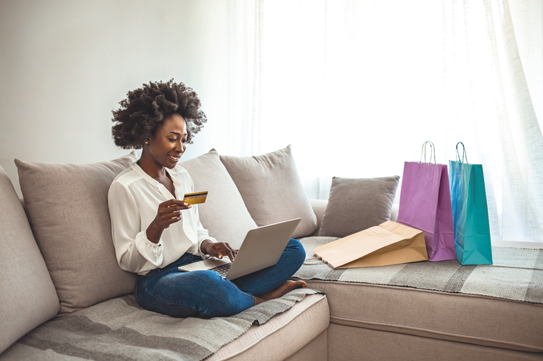 Fotografia. Uma mulher sentada em um sofá segurando um cartão de crédito em frente a um notebook. No sofá ao lado dela, há sacolas coloridas.
