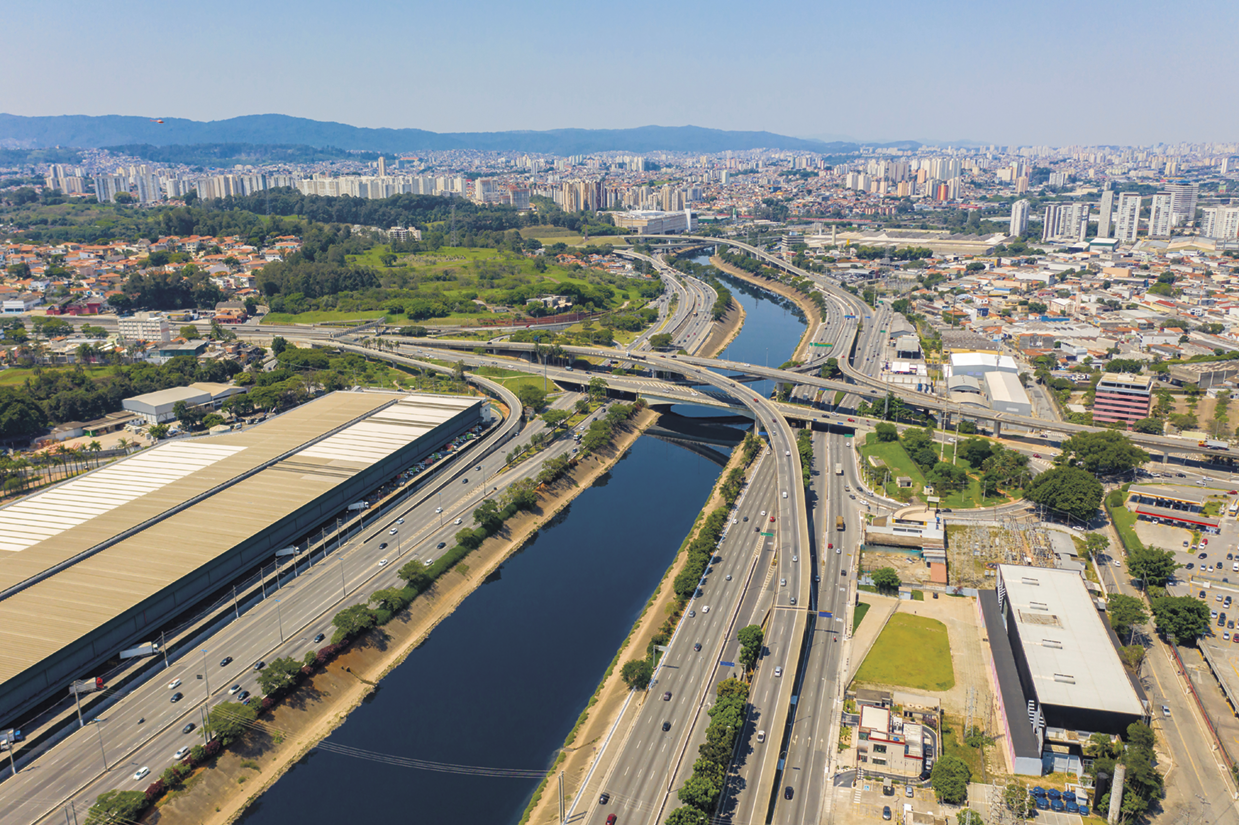 Fotografia. Vista de cima. Curso de um rio com avenidas nas laterais e pontes que o cruzam. Há áreas com vegetação, e construções nas margens.