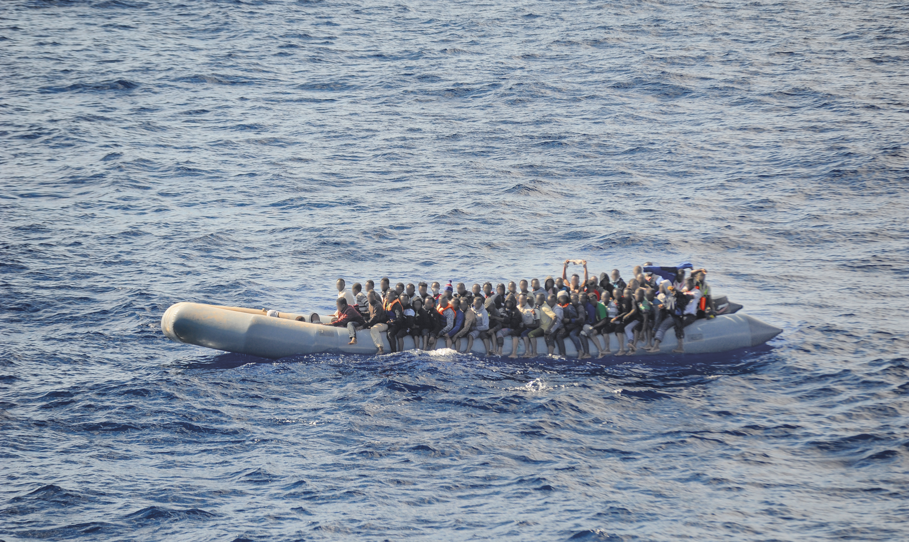 Fotografia. Um bote inflável com muitas pessoas agrupadas sobre ele, navegando no mar.