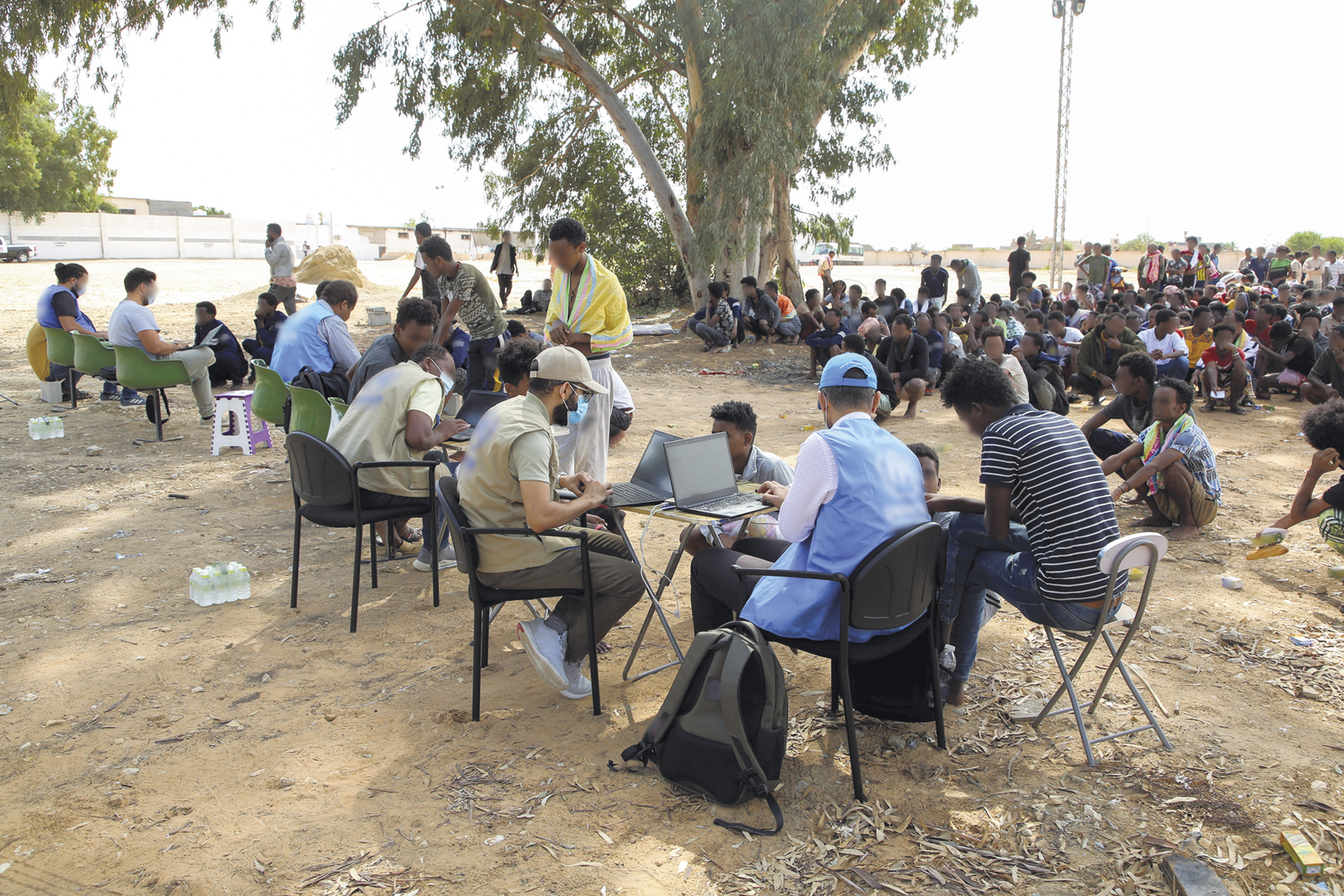 Fotografia. Pessoas reunidas em uma área aberta, com chão de terra. Algumas delas estão sentadas em mesas, com notebooks. E muitas estão reunidas ao fundo.