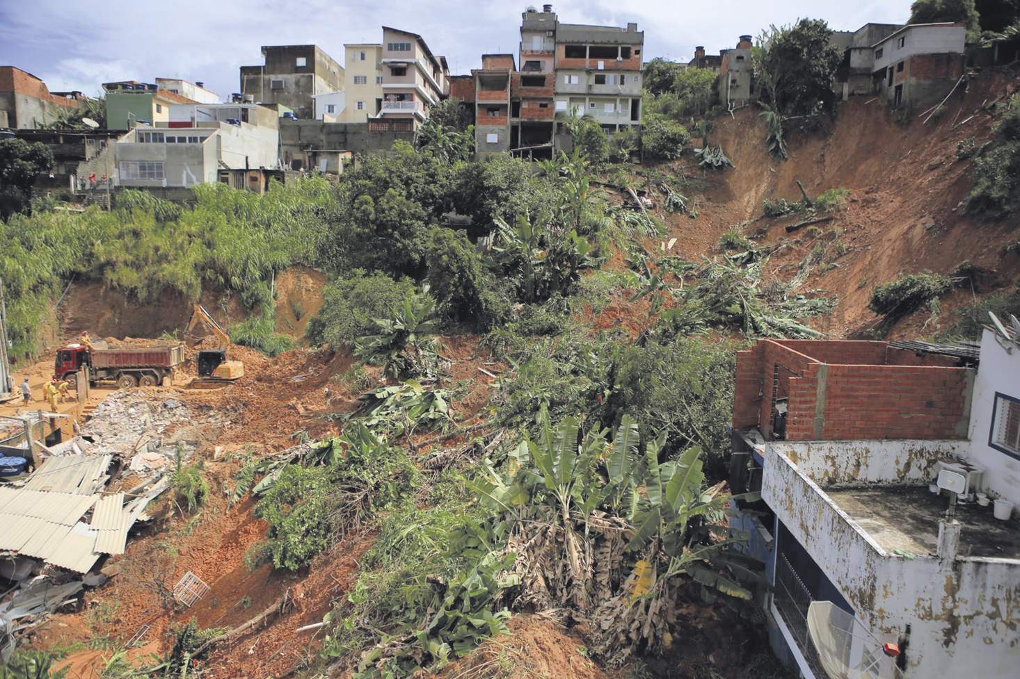 Fotografia. Deslizamento de um morro com árvores e destroços. Acima dele, construções.