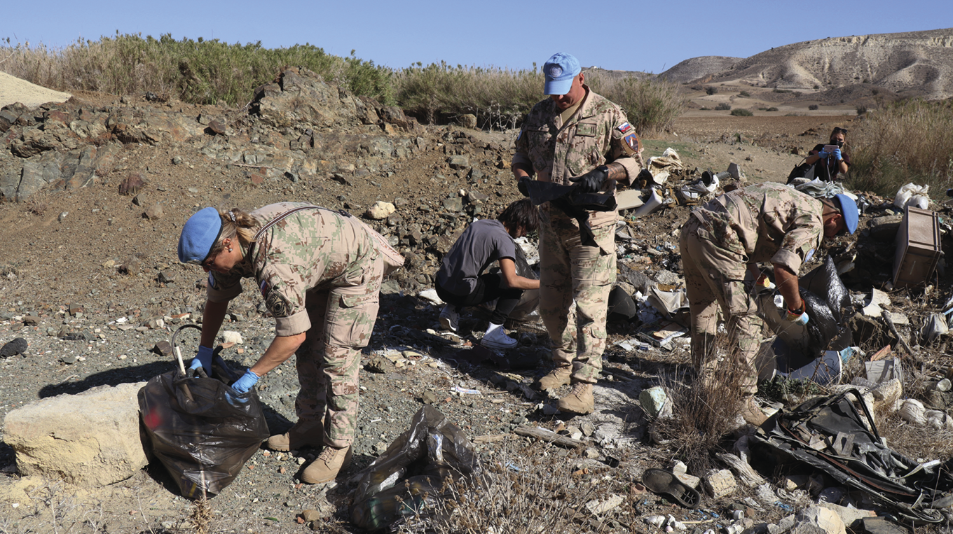 Fotografia. Soldados fardados em uma área de terra com lixo espalhado. Eles usam luvas e recolhem o lixo, segurando sacos pretos.