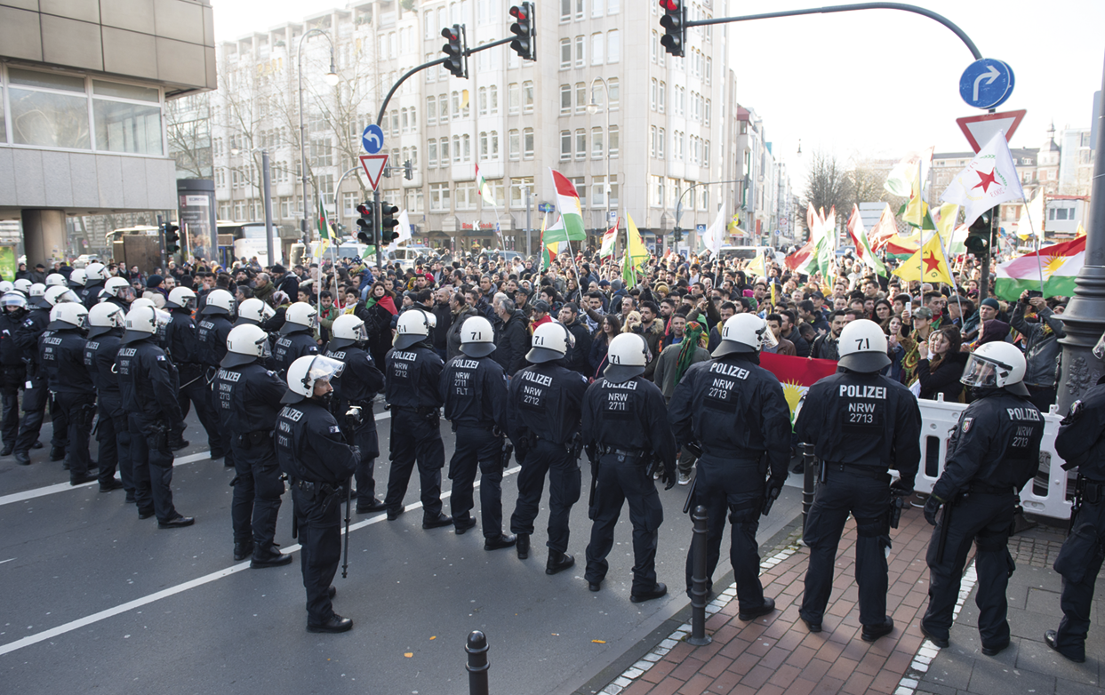 Fotografia. Policiais com uniformes e capacetes, parados enfileirados, em frente a uma multidão com bandeiras em uma rua. Ao fundo, edifícios.