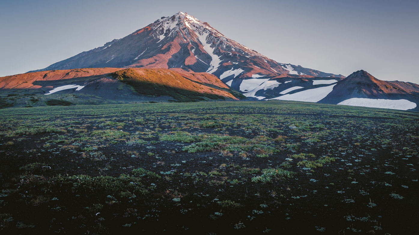 Fotografia. No primeiro plano, área plana com vegetação rasteira. Ao fundo, montanha com trechos cobertos por neve.