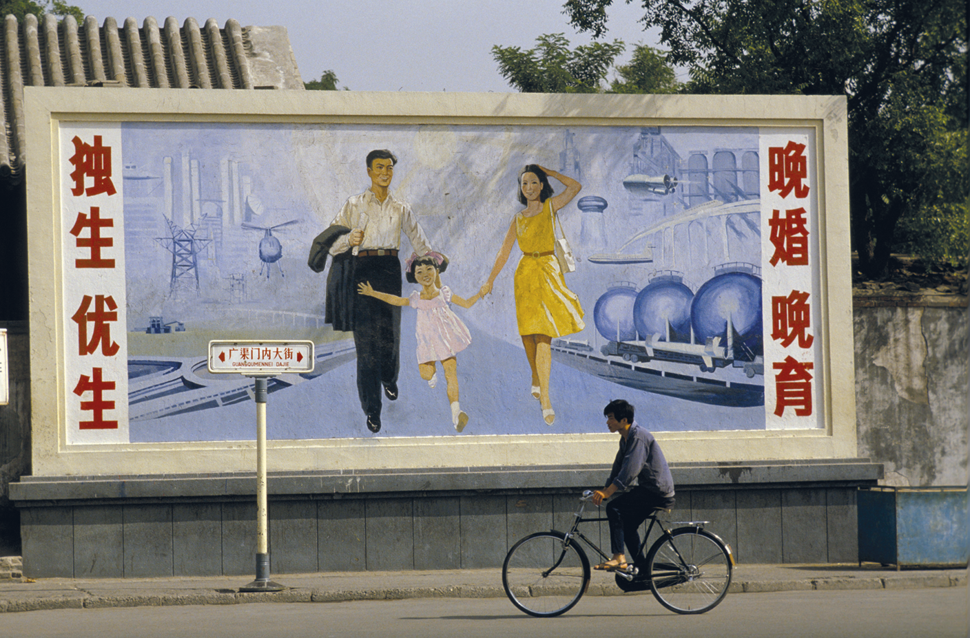 Fotografia. Um homem andando de bicicleta em frente a um painel com a figura de um homem e uma mulher caminhando com uma criança por uma rua, nas laterais há inscrições em chinês.