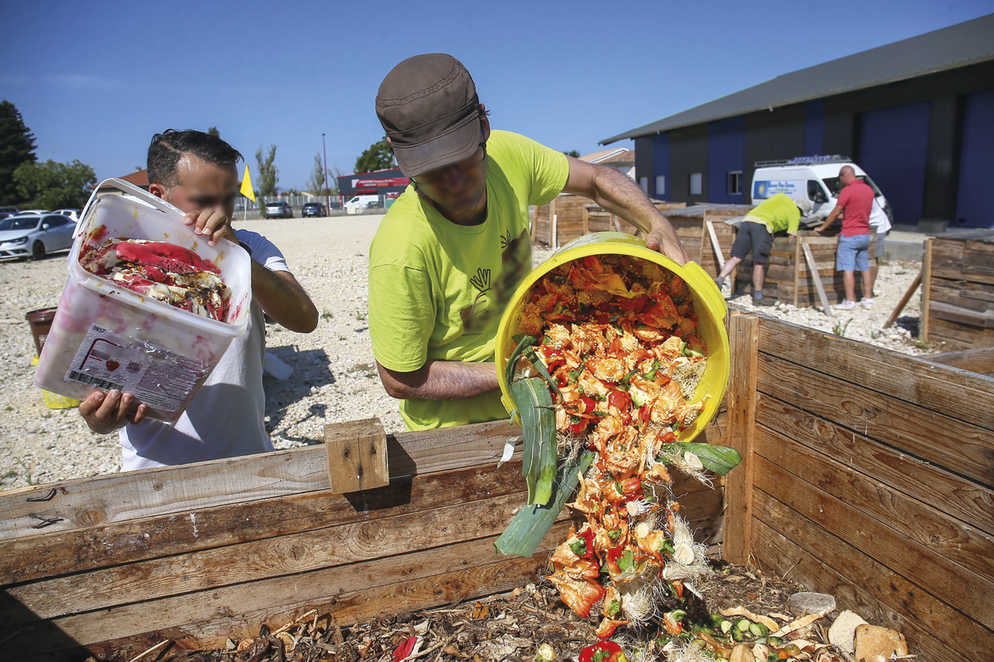 Fotografia. Dois homens despejando baldes de comida em uma lixeira. Ao fundo, construções.