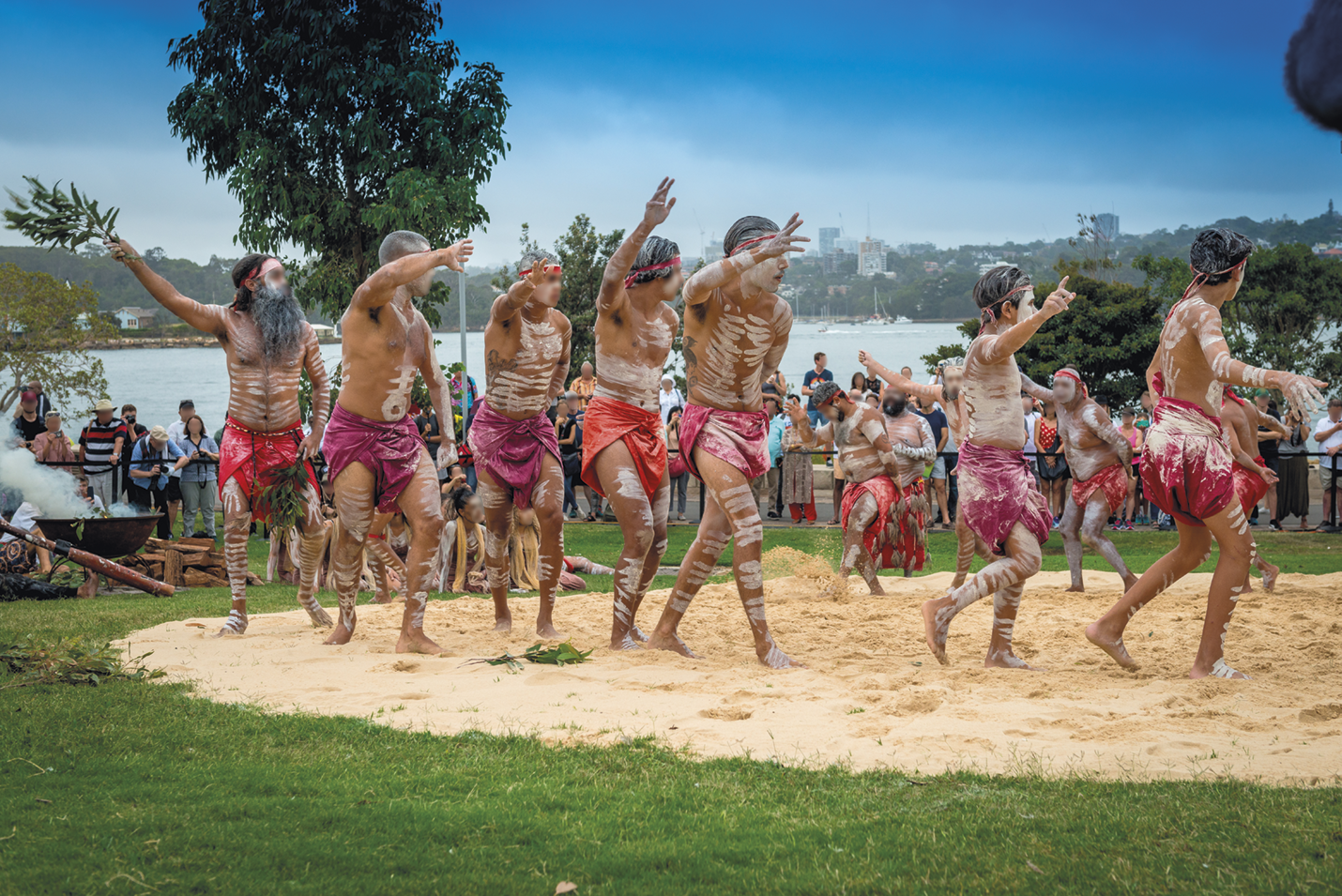 Fotografia. Grupo de pessoas com tecidos vermelhos e pinturas corporais espalhadas pelo corpo, dançando em uma área de areia cercada por grama. Ao redor, pessoas observando e árvores.
