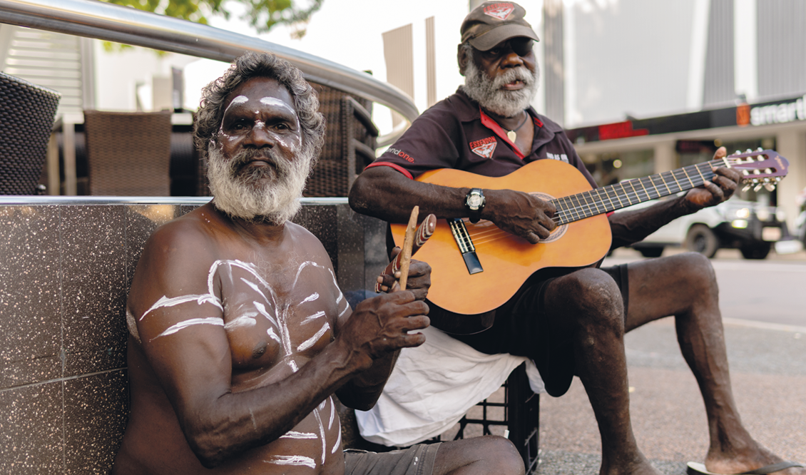 Fotografia. Dois homens sentados lado a lado, um deles toca violão e outro segura um objeto e possui pinturas corporais.