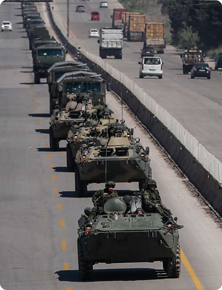 Fotografia. Veículos de guerra verdes trafegando enfileirados. Há soldados nos últimos carros. Nas laterais há carros e caminhões.