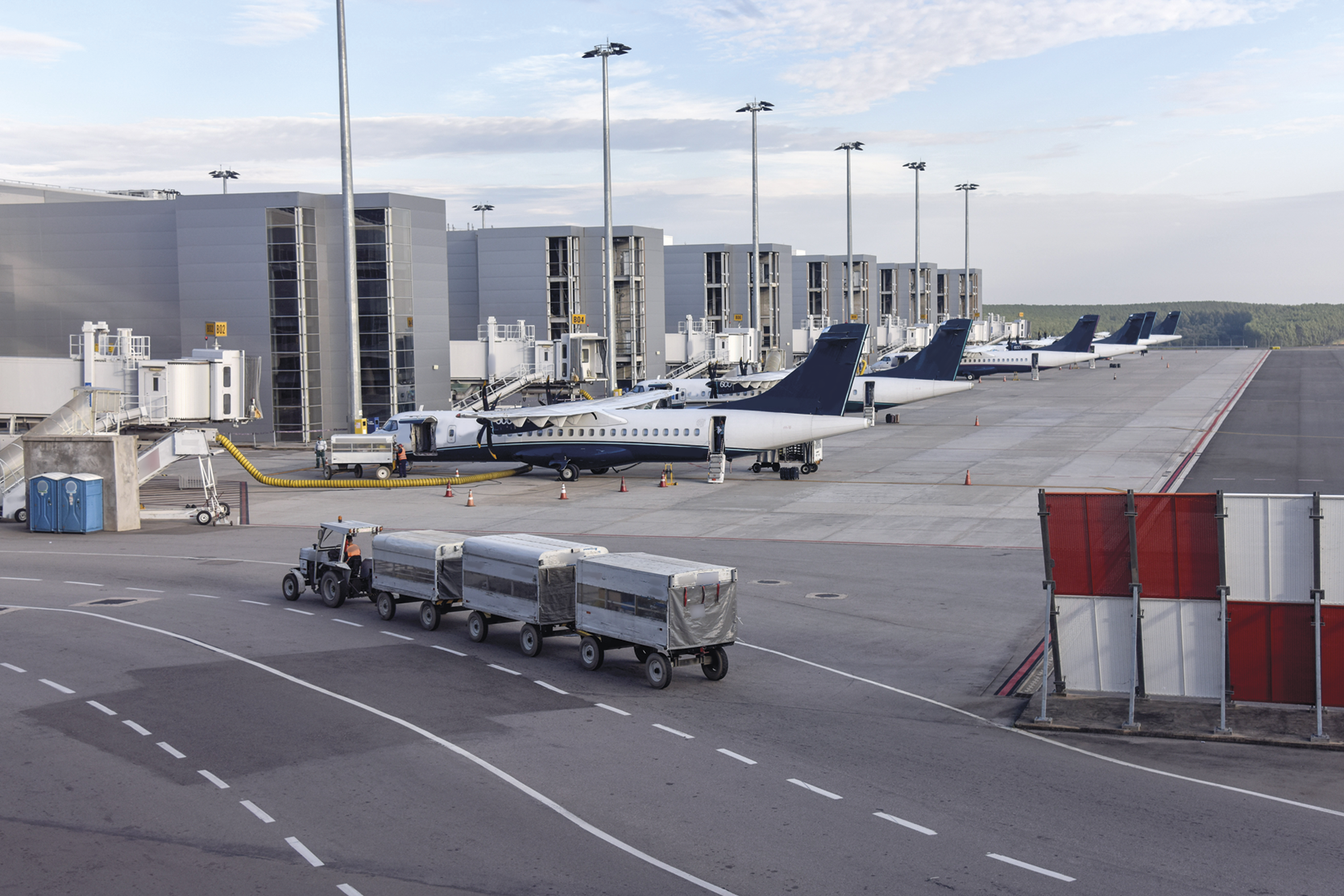 Fotografia. Aeroporto com aviões parados próximos a construções. Há um veículo acoplado a três engates malas na frente.