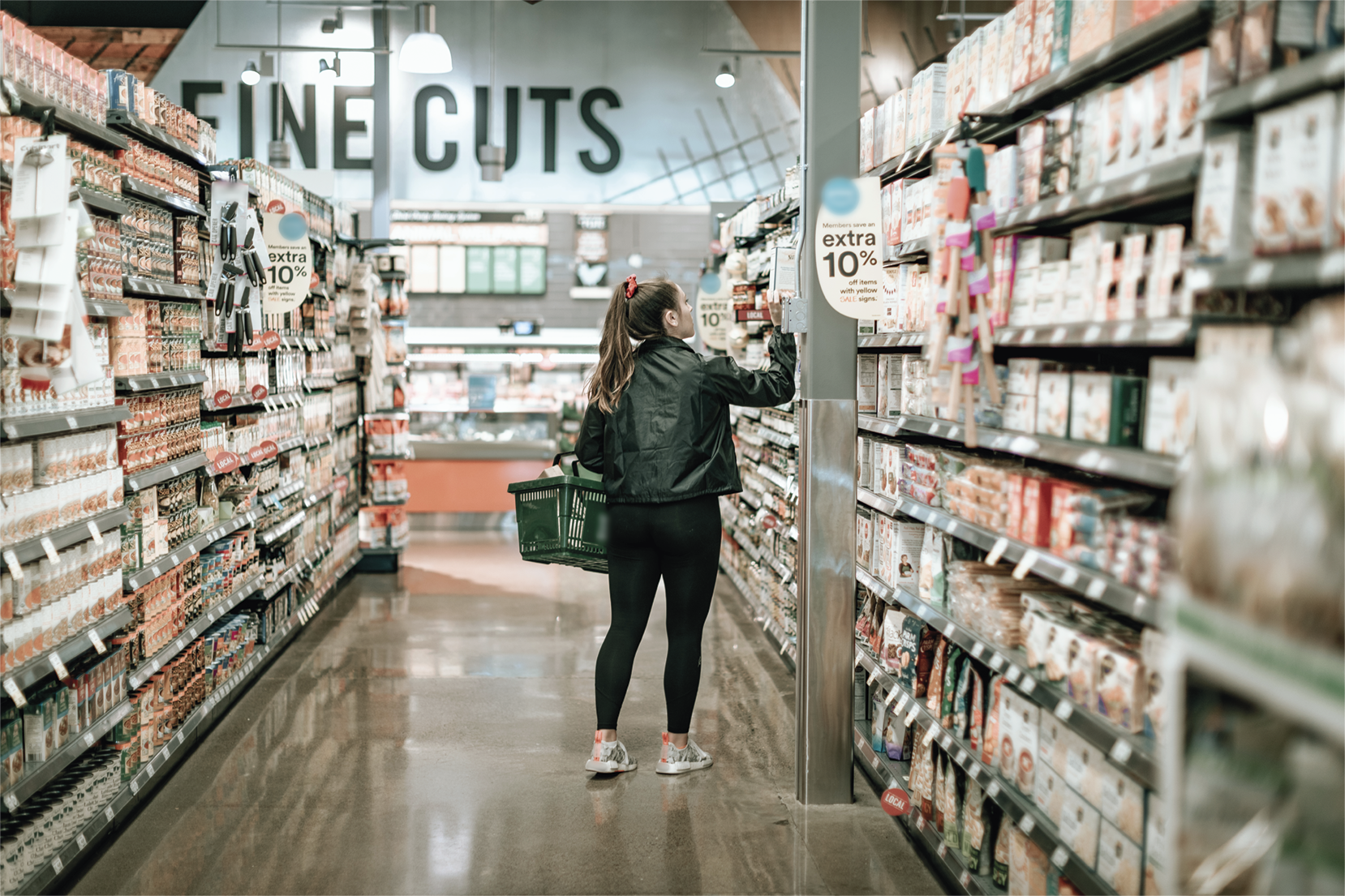Fotografia. Uma mulher caminhando pelo corredor de um supermercado, olhando um produto. Ela carrega uma cesta em um dos braços.
