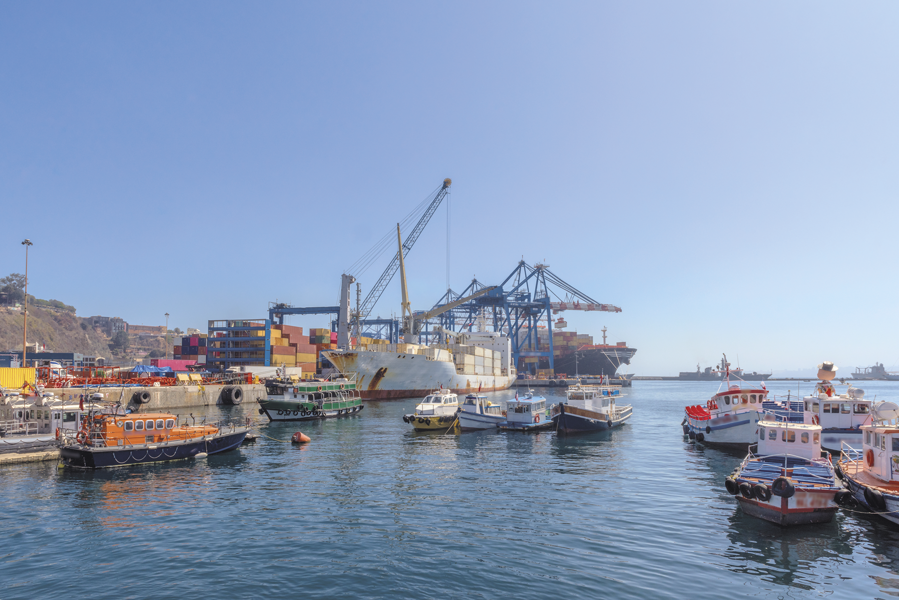 Fotografia. Um porto repleto de barcos e navios sendo carregados com contêineres.