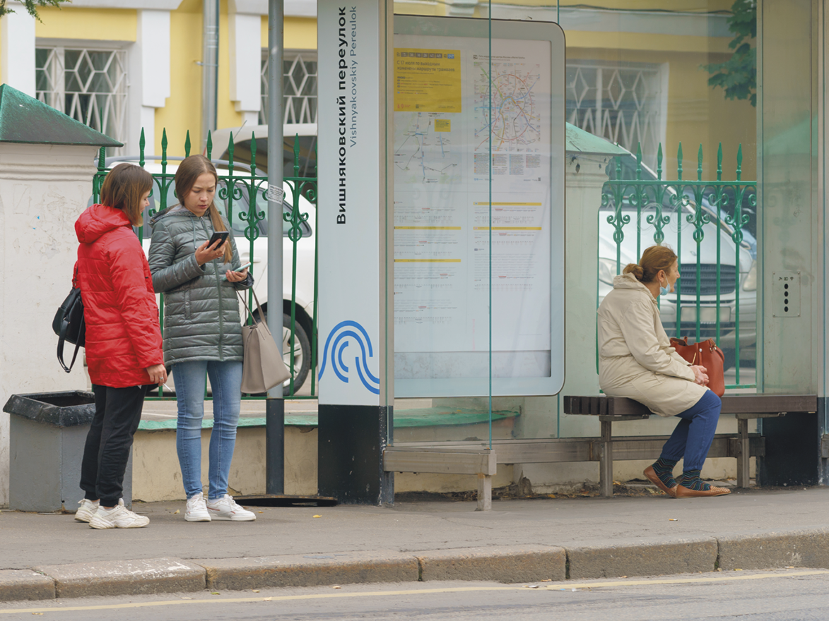 Fotografia. Duas meninas reunidas, uma delas segura dois celulares. Ao lado, uma pessoa em um ponto de ônibus.