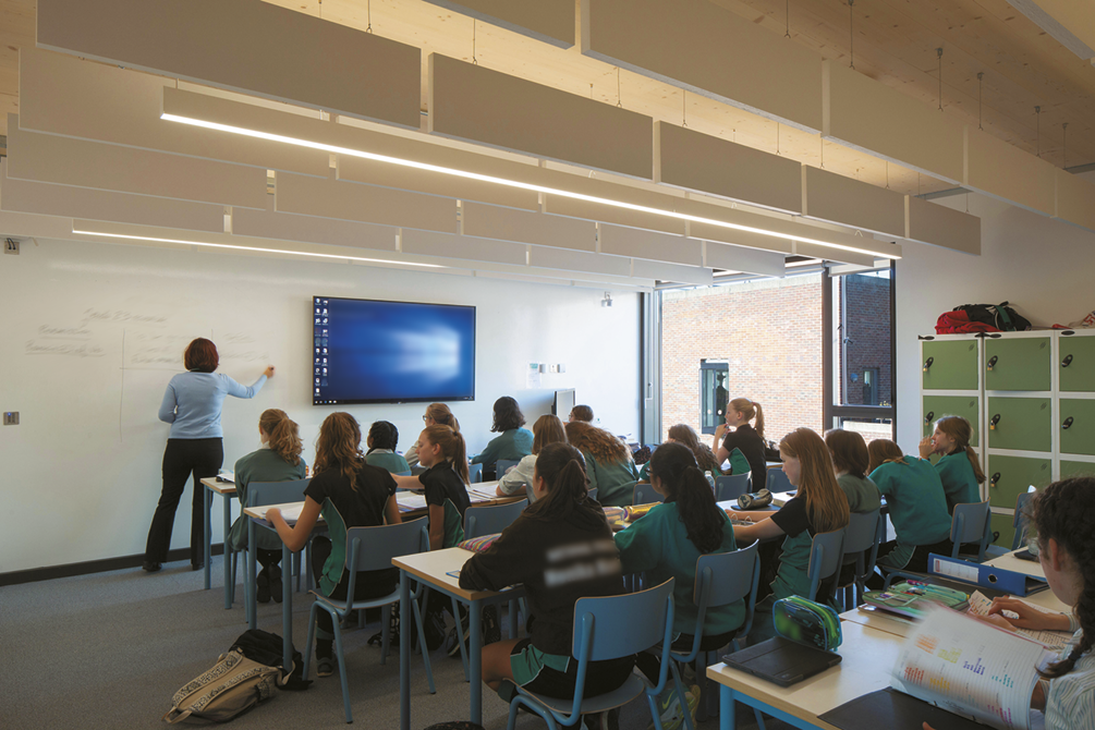 Fotografia. Sala de aula repleta de pessoas sentadas em frente a uma tela e uma mulher que escreve em uma lousa.