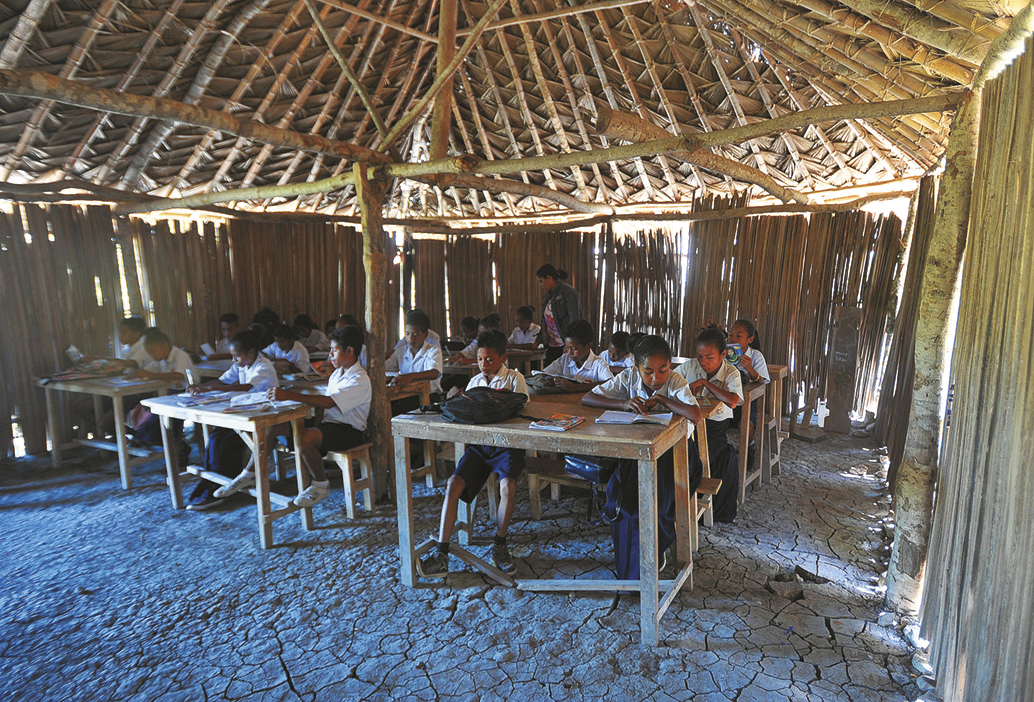Fotografia. Crianças sentadas em uma construção de tábua, com telhado de palha e chão de terra.