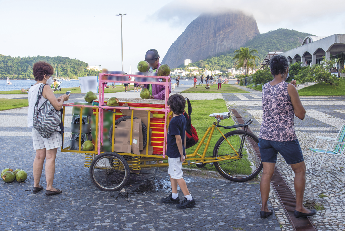 Fotografia. Um vendedor ambulante de água de coco, com um carrinho acoplado a uma bicicleta. Há algumas pessoas ao redor dele, e ao fundo, uma praia.