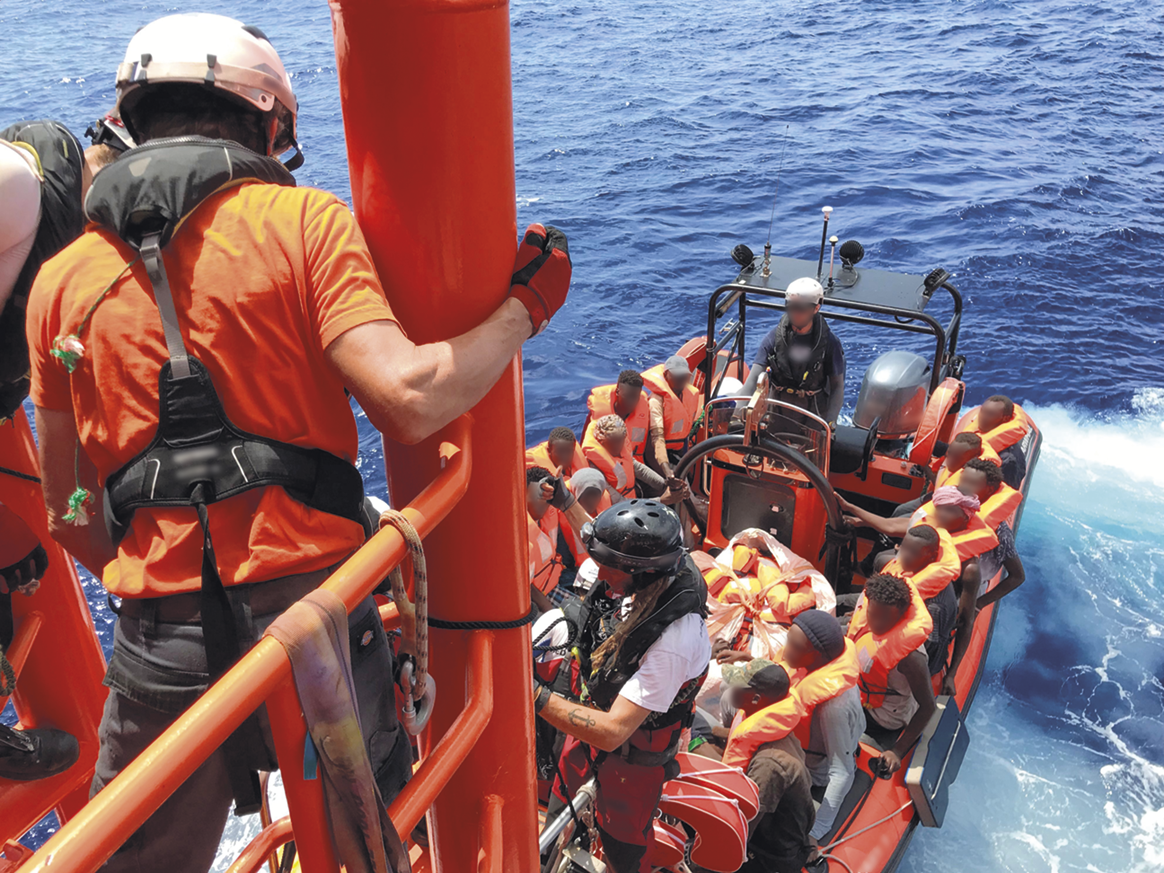 Fotografia. Vista de cima. Pessoas em um bote salva-vidas, usando coletes laranjas e capacetes brancos. Há uma pessoa se segurando em uma coluna em frente a ele.