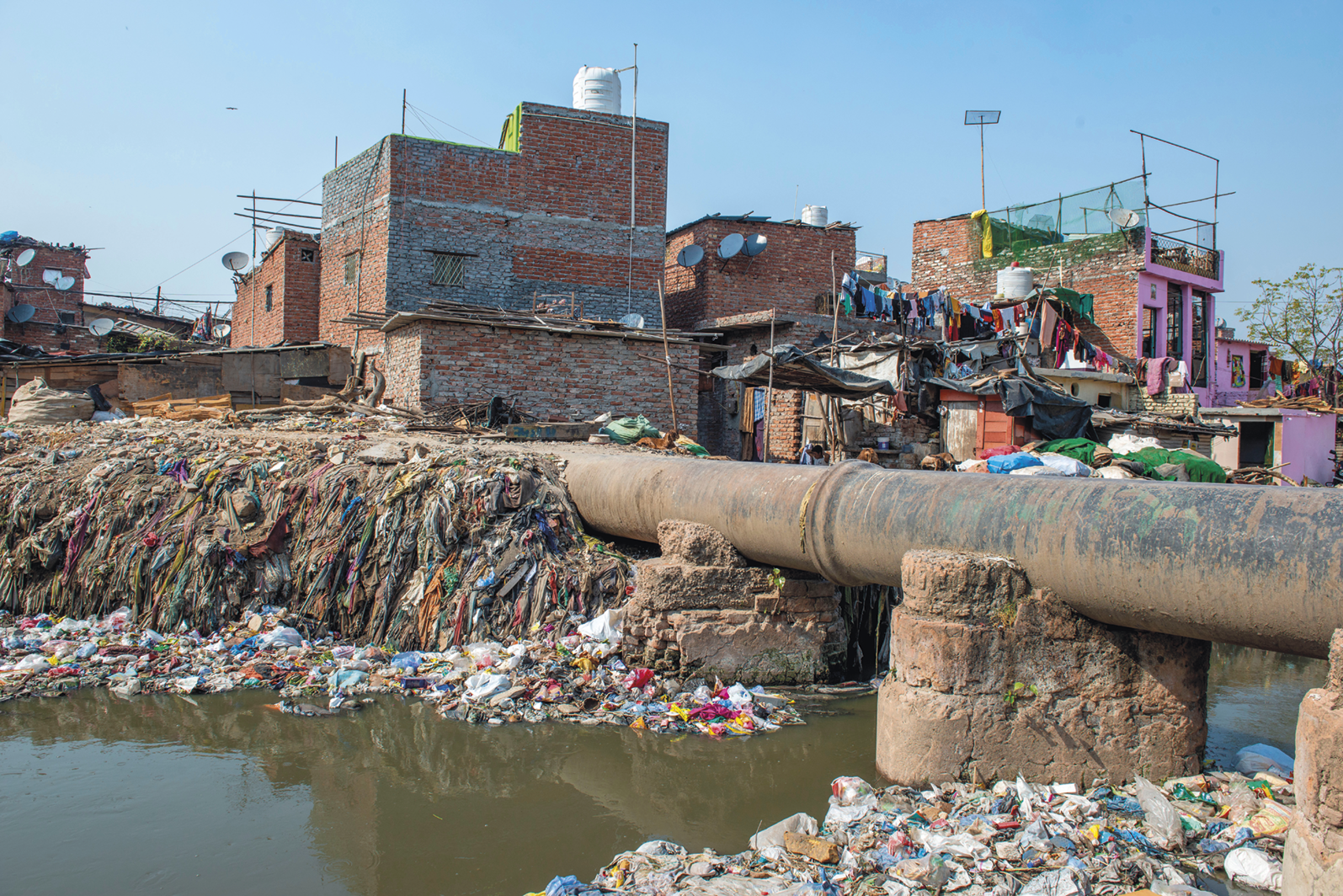 Fotografia. Rio repleto de lixo boiando e preso na margem. Há um duto que passa sobre o rio. Ao fundo, casas com tijolos expostos agrupadas.