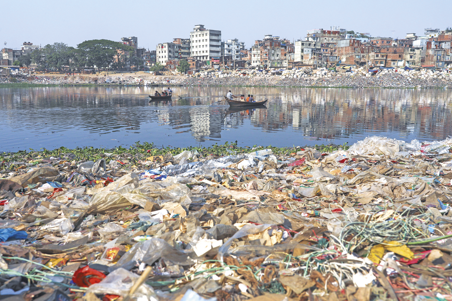 Fotografia. Margem de um rio repleto de lixo. Ao fundo, dois pequenos barcos e a outra margem com mais lixo, construções e árvores.