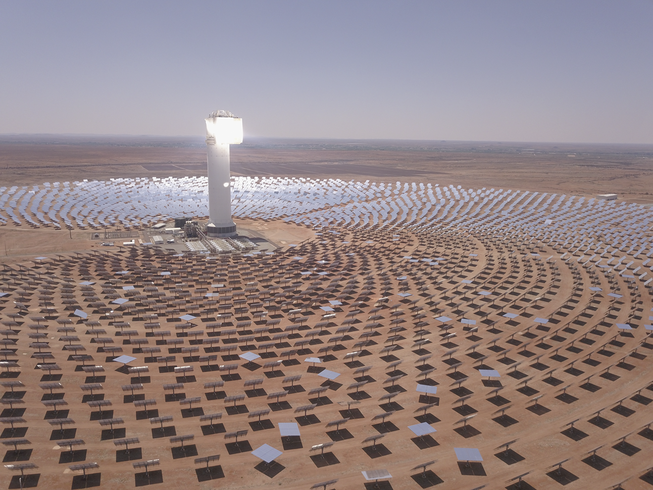 Fotografia. Uma grande quantidade de painéis solares dispostos em círculos ao redor de uma construção com uma torre iluminada no deserto.