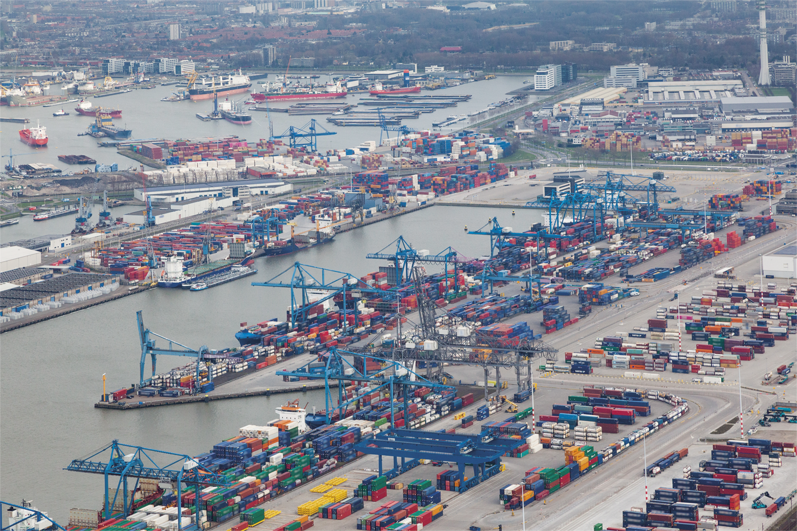 Fotografia. Vista do alto. Porto com diversos contêineres espalhados e navios ancorados ao fundo.