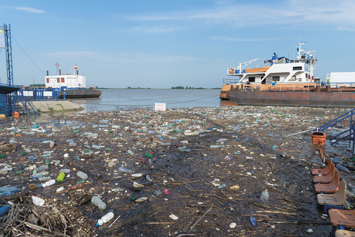 Fotografia. Rio coberto por lixo flutuando. Ao fundo, barcos ancorados.